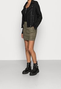 Veste de motard en suede noir avec des zippers argentés, jupe mini en suede verte olive, et bottines noires à boucles et semelles épaisses.