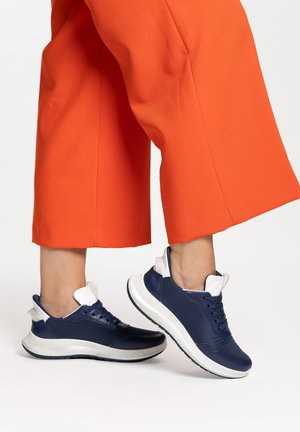 Füße in marineblauen und weißen Sneakers, kombiniert mit knöchellangen, hellorangefarbenen, weit geschnittenen Hosen auf weißem Hintergrund.