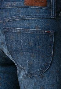 Denimjeans i mörkblått med stjärnmönster, har en bakficka och en läderetikett. Tyget har en strukturerad känsla.