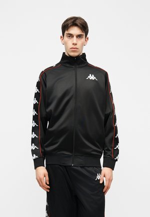 Mladi moški, oblečen v črno športno jakno z zadrgo in hlače, ki se ujemajo, z belimi logotipi na rokavih in nogavici, stoji pred enobarvnim ozadjem.