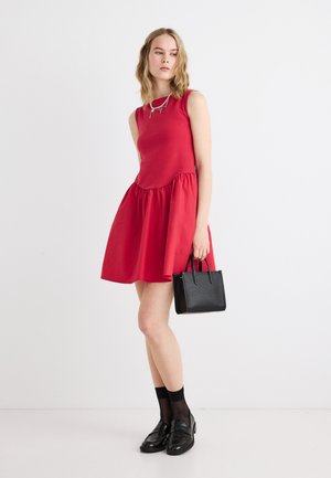 Donna con vestito rosso senza maniche, calzini neri alla caviglia, mocassini neri, che tiene una piccola borsa nera, posando su sfondo bianco.