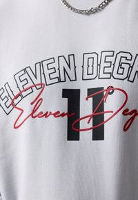 Camiseta blanca de algodón con un texto en negrita negro y rojo que dice "ELEVEN DEG", con el "11" destacado en negro y rojo con acentos en cursiva.