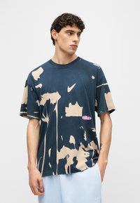 Νεαρός άνδρας που φοράει μπλε σκούρο και μπεζ μπλουζάκι Nike με εφέ βαφής και ανοιχτό μπλε σορτς Nike, στέκεται μπροστά σε λευκό, απλό φόντο.