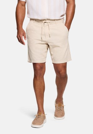 Hombre con pantalones cortos beige claros con cordón, zapatos casuales y camisa blanca de manga corta, de pie sobre un fondo liso.