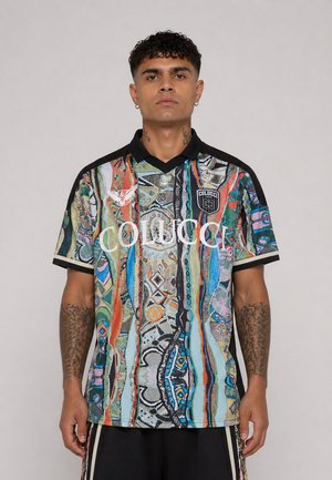 Man met tatoeages die een kort mouw, meerkleurig patroon overhemd draagt met de tekst "Colucci" en een zwarte kraag, staand tegen een effen achtergrond.