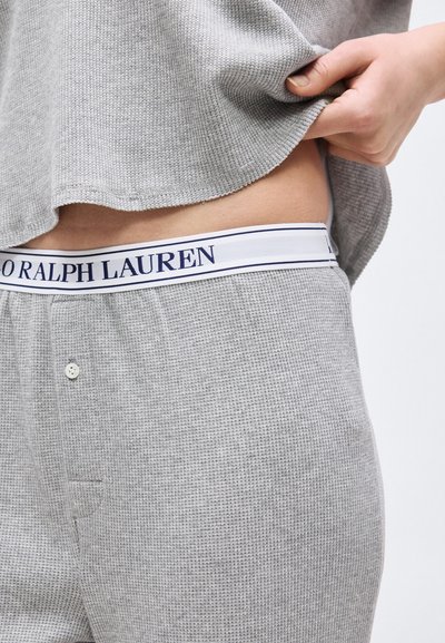 Άτομο που σηκώνει γκρι πουλόβερ πλεκτό, αποκαλύπτοντας ταιριαστό γκρι παντελόνι και λευκή ελαστική μέση με την επιγραφή «Polo Ralph Lauren».