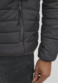 Veste matelassée grise avec une poche zippée, présentant un matelassage horizontal et des poignets élastiques. Texture lisse avec une coupe ajustée.