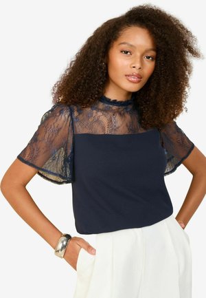 Haut bleu marine avec un corps uni et des manches courtes en dentelle transparente. Présente des motifs floraux et un col froncé. Associé à un pantalon de couleur claire.