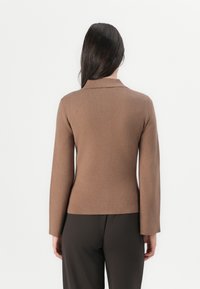 Maglione marrone aderente con maniche lunghe e colletto a punta. Texture liscia e design semplice, indossato sopra pantaloni scuri.