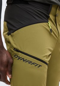 Κοντινό πλάνο σε ελαιοπράσινο παντελόνι Dynafit με μαύρες τσέπες με φερμουάρ και πράσινο φερμουάρ, φορεμένο από άτομο με σκούρο δέρμα.