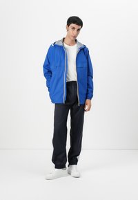 Jachetă albastră cu glugă gri, cu fermoar la față și două buzunare pe piept, asortată cu pantaloni negri și adidași albi.