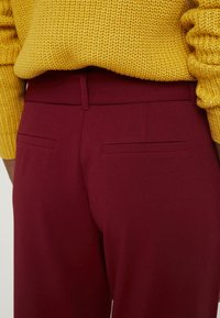 Pantalons de costume bordeaux avec poches arrière plates, associés à un pull en maille jaune confortable avec un design texturé et des poignets côtelés.