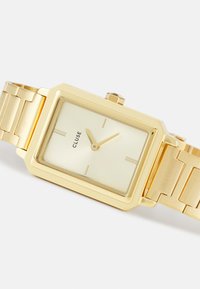 Montre en métal doré carré avec une finition brillante, un cadran léger doré texturé et des marqueurs d'heure minimalistes. Dotée d'un bracelet en maillons dorés.