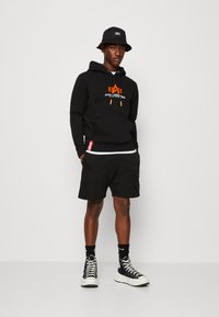 Sudadera con capucha negra y logotipo naranja, pantalones cortos negros y sombrero de cubo negro. Usada con zapatillas altas y calcetas de marca visibles.