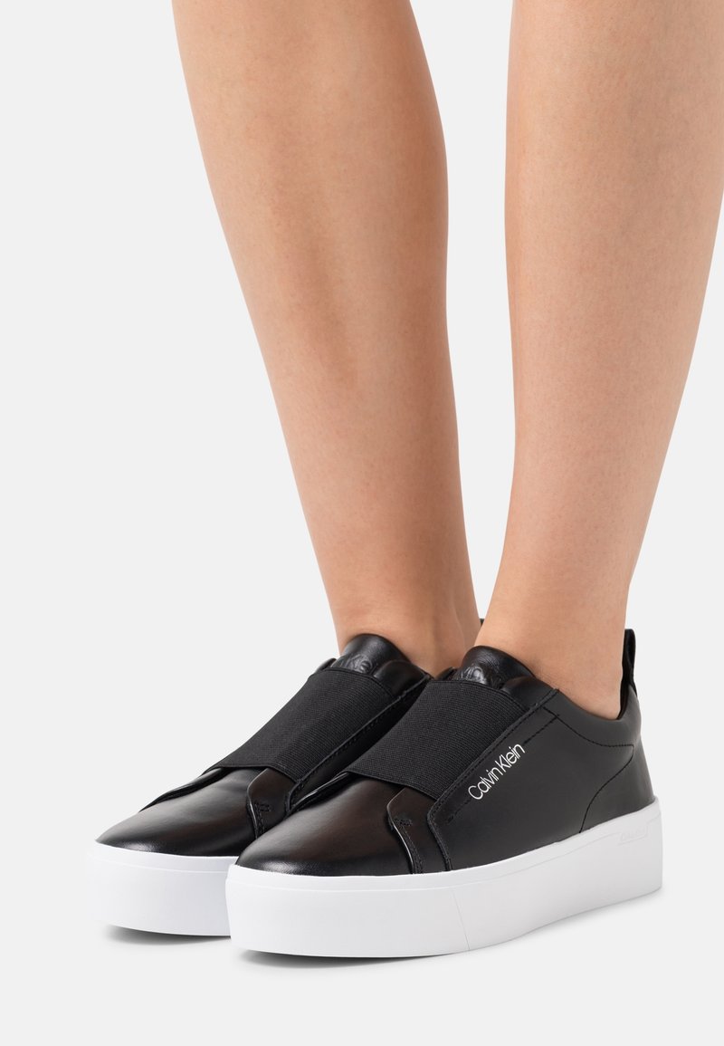 Czarne skórzane sneakersy typu slip-on z elastycznymi paskami i białymi platformami, z subtelnym detalem logo z boku. Gładka faktura.