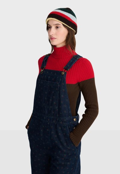 Jeune femme portant un pull côtelé rouge et marron, une salopette en jean foncé à motifs et un bonnet tricoté rayé, debout les mains dans les poches.