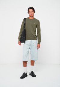 Man in een olijfgroen trui, lichtblauwe lange denim shorts, witte sokken, zwarte loafers en met een zwarte schoudertas staat tegen een witte achtergrond.