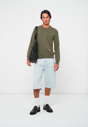 Mann iført olivengrønn genser, lyseblå lange denimshorts, hvite sokker, sorte loafers og bærende en svart skulderveske står mot en hvit bakgrunn.