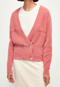 Femme portant un cardigan en peluche rose doux avec des boutons dorés sur un haut côtelé blanc et une jupe crème, vue des épaules aux hanches.