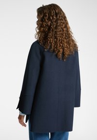 Cappotto blu navy con una silhouette rilassata, maniche lunghe e una finitura leggermente texturizzata. La vista posteriore mostra un design liscio con minimi dettagli.