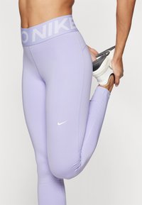 Leggings lila cu o talie lată, având inscripția "NIKE" în alb și un mic logo swoosh alb. Realizate dintr-un material neted și elastic.