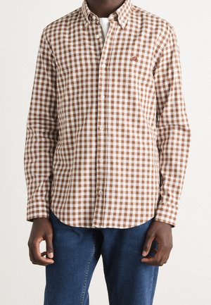 Hombre con una camisa de cuadros marrón y blanca abotonada sobre una camiseta blanca y jeans azules, de pie frente a un fondo liso.