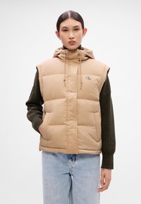 Beige vzdušná vesta s kapucí, se zapínáním na zip, předními kapsami a logem. Nosit over tmavě zeleným dlouhým rukávem.