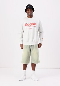 Γκρίζα φούτερ με κόκκινη εκτύπωση "Kodak" και "Huf", συνδυασμένη με ανοιχτό πράσινο cargo σορτς. Μαύρο καπέλο bucket και λευκά αθλητικά παπούτσια ολοκληρώνουν το ντύσιμο.