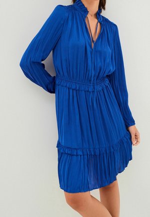 Femme portant une robe plissée bleue mi-longue à manches longues, avec un col à volants et une ceinture à la taille, sur un fond uni.