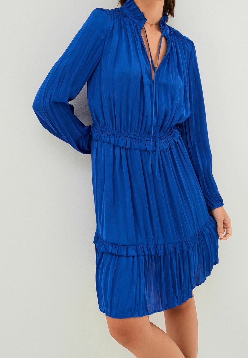 Femme portant une robe plissée bleue mi-longue à manches longues, avec un col à volants et une ceinture à la taille, sur un fond uni.