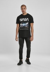 Černé bavlněné tričko s grafikou NASA, krátké rukávy a kulatý výstřih. Oblečeno s tmavě šedými džíny a černými teniskami.