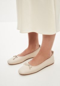 Ballerine in pelle beige con punta arrotondata, caratterizzate da un dettaglio a fiocco decorativo e un design minimalista a profilo basso e senza cuciture. Texture morbida, stile essenziale.