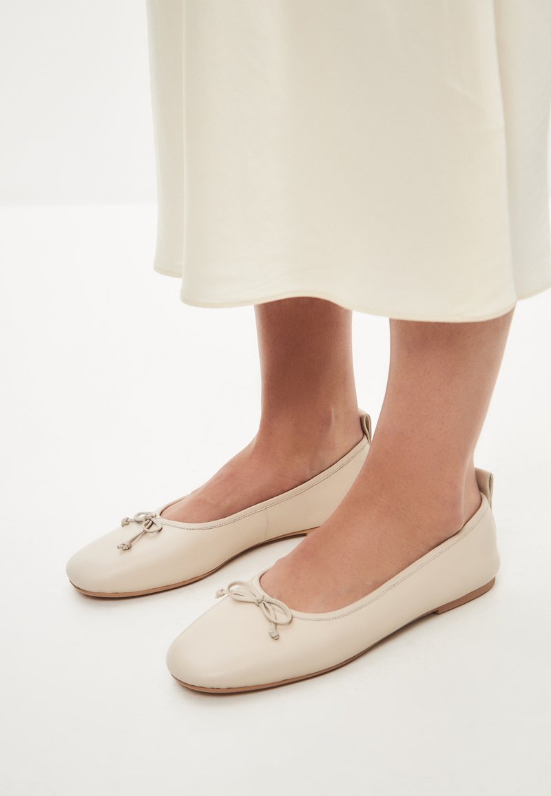 Ballerine in pelle beige con punta arrotondata, caratterizzate da un dettaglio a fiocco decorativo e un design minimalista a profilo basso e senza cuciture. Texture morbida, stile essenziale.