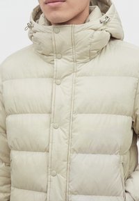 Homme portant une veste matelassée beige clair avec col montant, fermeture par boutons, poches zippées et capuche ajustable avec cordons.