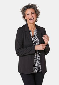 Zwarte blazer met een maatwerk ontwerp, voorzien van twee voorzakken, gedragen over een wit met zwart patroon blouse. Glad stof, gestructureerde vorm.