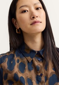 Woman with long dark hair wearing a blue and brown animal print shirt and square gold earrings, looking slightly upward.