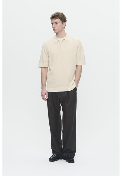 Polo en maille beige clair, à manches courtes et col, associé à un pantalon sombre ample et des chaussures noires. Design sobre.