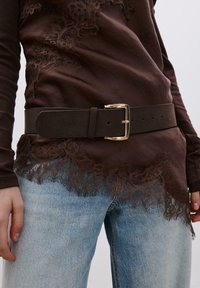 Hnedý látkový top s čipkovanou lemovkou, doplnený širokým tmavým opaskom so zlatou prackou. Svetlomodré džínsové nohavice s voľným strihom.