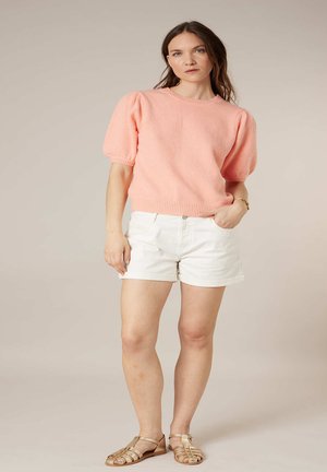 Femme debout portant un pull à manches courtes rose, un short blanc, des sandales dorées et un bracelet sur un fond beige.