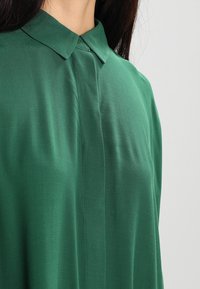 Blusa verde con colletto realizzata in un tessuto leggero e morbido. Presenta un design pulito con una patta frontale e una consistenza lieve.