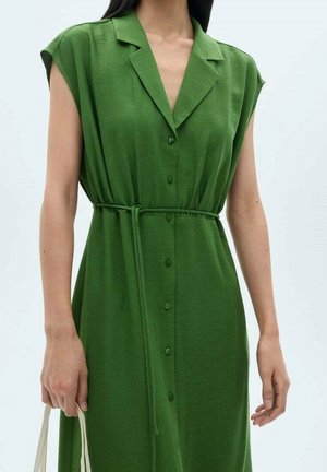 Femme portant une robe verte sans manches à boutons avec un col et une ceinture nouée, tenant les poignées d'un sac fourre-tout beige dans sa main gauche.