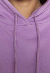 Lila Kapuzenpullover mit einer Kordelzugkapuze, glatter Stoffoberfläche und geflochtenen Kapuzenbändern. Sichtbare Steppnähte entlang des Halsausschnitts.