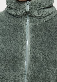 Zielona polarowa kurtka z wysokim kołnierzem, z suwakiem z przodu. Materiał ma miękką, teksturowaną powierzchnię z puszystym wykończeniem.