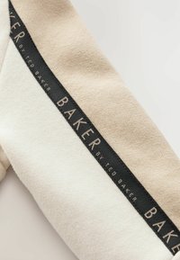 Tissu bicolore beige et crème avec une étiquette noire portant "BAKER par Ted Baker", présentant une surface texturée et des lignes épurées.