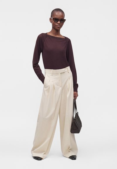 Femme aux cheveux courts portant un haut à manches longues sombre, un pantalon large crème, des chaussures noires, des lunettes de soleil et tenant un petit sac à main noir.