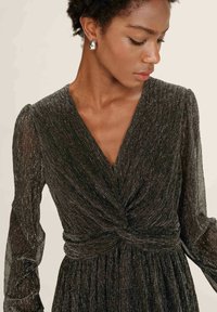 Donna con capelli corti e ricci che indossa orecchini a forma di goccia d'argento e un vestito metallizzato nero a maniche lunghe con dettagli plissettati e nodo twisted.