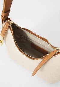 Beige canvas handbag with brown leather strap, gold-tone zipper, and interior pocket; smooth texture and logo detail visible.