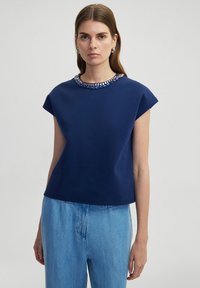 Marineblauwe top met korte mouwen en een ronde halslijn, versierd met een decoratieve kralen kraag. Heeft een ontspannen pasvorm en een zachte stoftextuur.