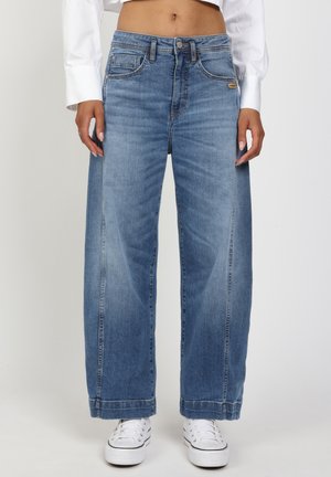 Blaue Jeans mit mittlerer Taille und weitem Bein, getragen mit weißen Turnschuhen und einem weißen, kurz geschnittenen Langarmshirt, die Hände entspannt an den Seiten.