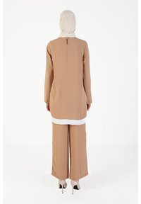 Tunique beige à manches longues, ourlet blanc, découpe au dos, associée à un pantalon beige à jambes larges et des chaussures à talons hauts. Texture de tissu lisse.